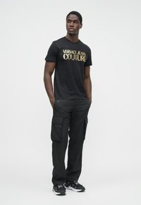 Vīrietis, kas ģērbies melnā Versace Jeans Couture T-kreklā ar zeltītām burtiem, melnās cargo biksēs un melnās sporta kurpēs, stāvot ar rokām kabatās.