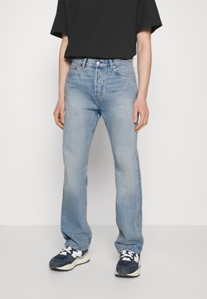 Personne portant un jean droit bleu clair, un t-shirt noir et des baskets noires et blanches, debout devant un fond blanc uni.