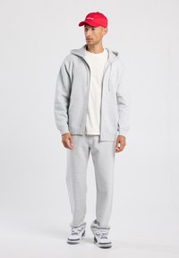 Sudadera con cremallera de color gris claro con cordones, combinada con pantalones de chándal a juego. Camiseta blanca por debajo y gorra de béisbol roja. Tejido texturizado.