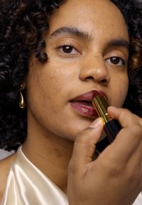 Un rouge à lèvres avec un capuchon doré est appliqué sur les lèvres d'un mannequin, présentant une teinte rouge profond. Le mannequin a des cheveux bouclés et porte un haut en satin clair.