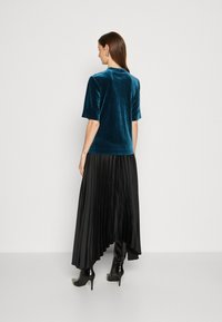 Teal fluwelen top met korte mouwen en een gladde textuur, gecombineerd met een zwarte plissé rok en zwarte hoge hakken. Eenvoudige halslijn en manchetten.