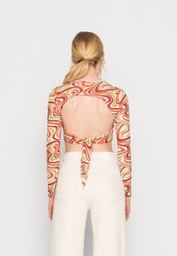 Blusa de mangas longas com um padrão espiral em vermelho, verde e creme. Apresenta uma costa aberta e um detalhe amarrado na cintura. Combinada com calças brancas de cintura alta.