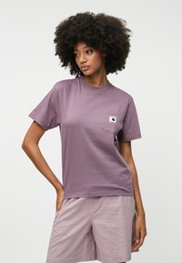 Λιλά κοντομάνικο μπλουζάκι με τσέπη στο στήθος και λογότυπο, φτιαγμένο από μαλακό και λείο ύφασμα, συνδυασμένο με υφασμάτινα ανοιχτό ροζ σορτς.