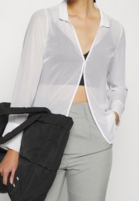 Camisa blanca ligera y transparente con cuello, mangas largas y ribete en contraste. Combinada con un bolso negro acolchado y pantalones grises.