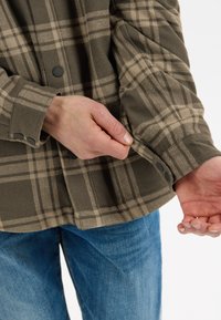Bruin en beige geruite flanellen shirt met zwarte knopen, met een knoopmanchet en een ontspannen pasvorm. Lichte blauwe spijkerbroek zichtbaar eronder.