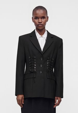 Femme aux cheveux courts portant un blazer noir à boutons sur le devant et un laçage style corset sur une chemise blanche rayée.