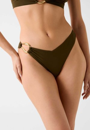 Kobieta w oliwkowo-zielonych majtkach od bikini z złotym metalowym pierścieniem na biodrze, pokazująca środkową część ciała i górną część ud na jednolitym tle.