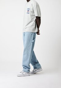 Harmaa T-paita, jossa on sinistä tekstiä ja logo, yhdistettynä vaaleansinisiin löysiin farkkuihin ja harmaisiin Nike-tossuihin, joissa on valkoisia yksityiskohtia.