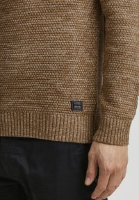 Pull marron tricoté avec un motif texturé, ourlet et poignets côtelés. Il présente une petite étiquette noire près du bas avec les informations de la marque.