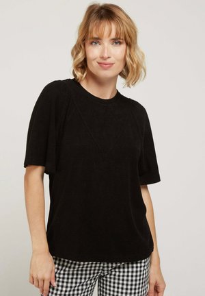 Haut noir à manches courtes avec un col rond, présentant une finition texturée et un subtil détail de couture diagonale. Arrière-plan minimal, pantalon à carreaux visible.