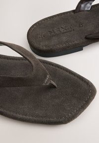 Brune flip-flops i ruskind med en flad sål, der har en V-formet rem og diskrete syninger langs kanterne, som viser et minimalistisk design.