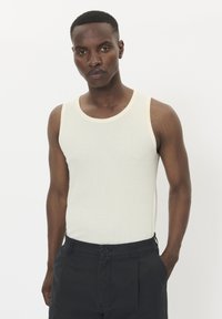 Világos krém színű, bordázott tank top, krém színű díszítéssel. Sötét tengerészkék rövidnadrággal párosítva. Az egyén semleges arckifejezéssel áll.