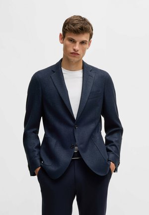 Blazer navy in tessuto strutturato, con rever a punta, due bottoni e una tasca sul petto, abbinato a una t-shirt bianca sotto.