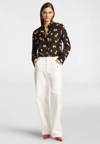 Blusa nera con stampa floreale, pantaloni bianchi a vita alta e décolleté rossi a punta. La modella sta in piedi con le mani in tasca, rivolta in avanti.