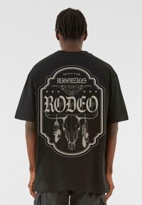 Svart t-skjorte med kort erm som har et stort grafisk trykk av et hodeskalle og fjær, med teksten "RODEO" tydelig vist.