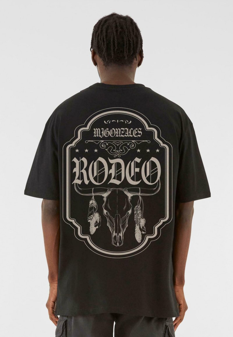 Svart t-skjorte med kort erm som har et stort grafisk trykk av et hodeskalle og fjær, med teksten "RODEO" tydelig vist.