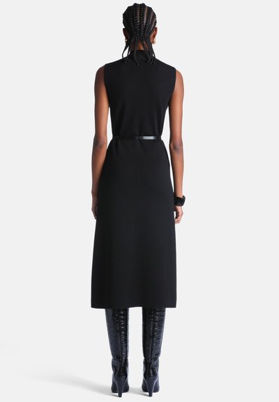 Femme aux cheveux tressés portant une robe noire sans manches, une ceinture noire et des bottes noires texturées jusqu'aux genoux, se tenant de dos sur un fond blanc.