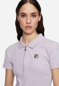 Lichtpaarse geribde polo met een rits aan de voorkant, korte mouwen en een ronde logo patch met rood, wit en blauwe accenten op de linkerborst.