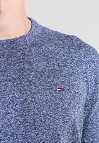 Blauer, strukturierter Pullover mit Rundhalsausschnitt, der ein kleines Logo in Rot, Weiß und Blau auf der linken Brust zeigt.