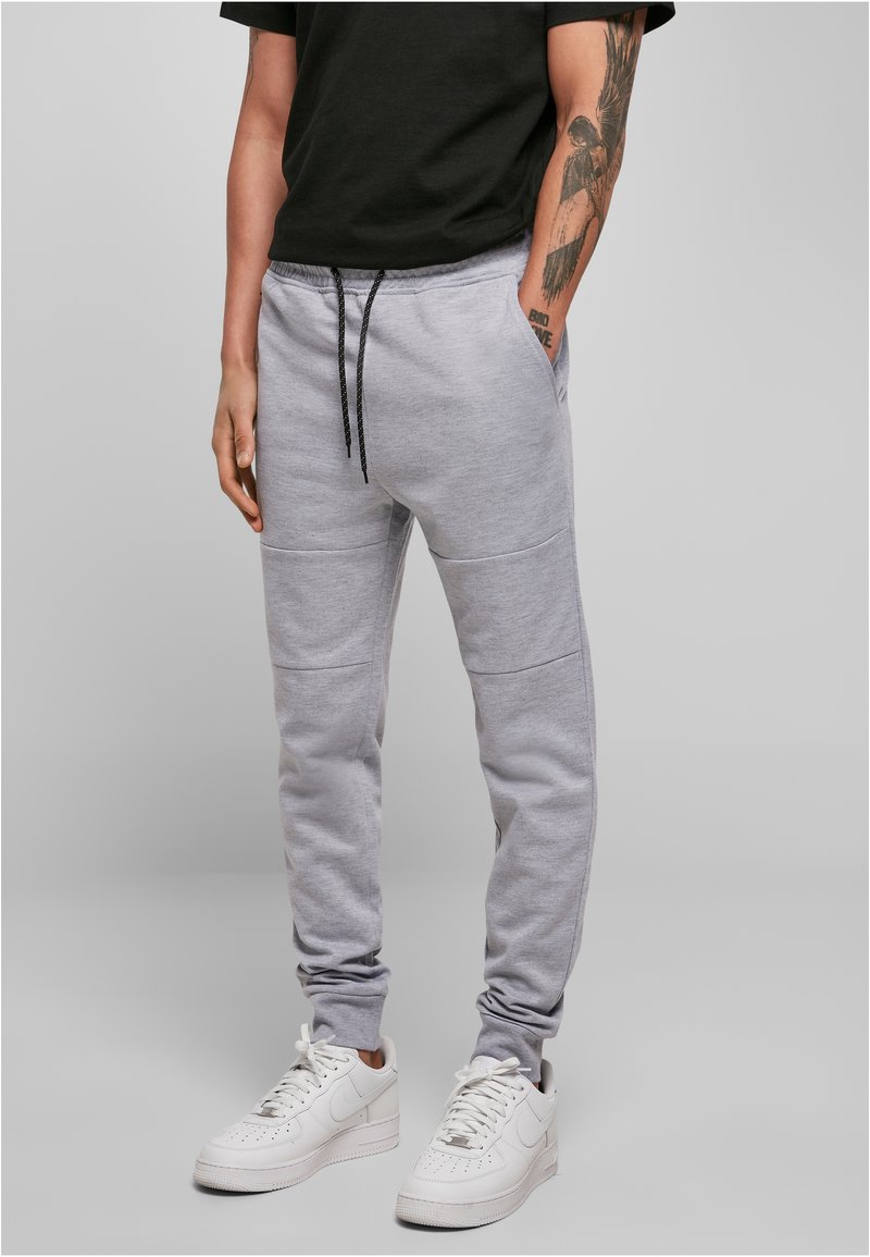 Pantalons de survêtement gris en matériau doux, avec poches latérales, taille élastique avec cordon de serrage et jambes fuselées. Associés à des baskets blanches.
