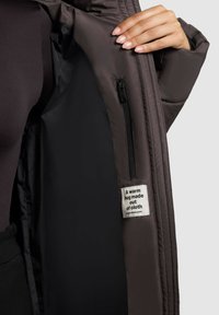 Unutrašnjost smešnog smešaka pufastog smešaka smešnog smešaka sa glatkim, tamnim podstavljem. Sadrži mali džep sa rajsferšlusom i oznaku koja glasi: "Topli zagrljaj napravljen od tkanine."