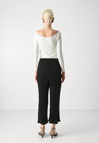 Femme aux cheveux blonds en chignon, portant un haut blanc à manches longues, un pantalon large noir et des talons ouverts en peau de serpent, vue de dos.