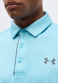 Κοντινό πλάνο ενός ατόμου που φοράει ανοιχτόγαλανη πόλο μπλούζα με κουμπιά και γκρι λογότυπο Under Armour στο στήθος.