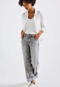 Donna con capelli ricci che indossa una camicia bianca aperta sopra una maglietta bianca, jeans grigi con risvolto, scarpe da ginnastica bianche, in piedi con le mani nelle tasche.