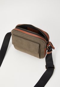 Sac bandoulière en toile vert olive avec des accents en cuir marron, fermeture éclair et poche avant ; sangle noire adjustable.