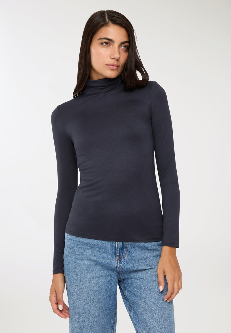 Młoda kobieta z długimi ciemnymi włosami, ubrana w czarny sweter z wysokim kołnierzem i niebieskie dżinsy, stojąca na tle jednolitego jasnego tła.