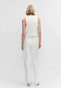 Kobieta z blond włosami do ramion, ubrana w bezrękawnik w kolorze białym, białe dopasowane spodnie i białe szpilki, stojąca na tle jednolitego tła.