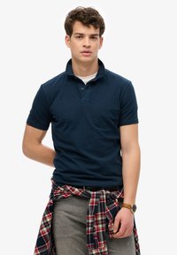Polo shirt blu navy con maniche corte e colletto, indossato sopra una t-shirt bianca. Jeans grigi e una camicia a quadri rossa annodata in vita.