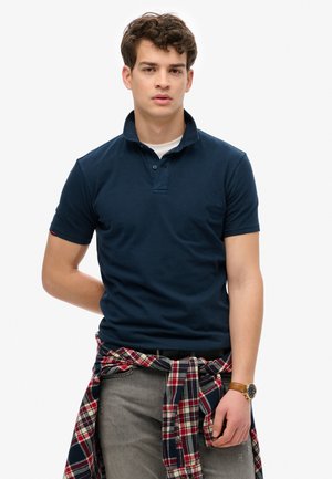Polo azul marino de manga corta y cuello, llevado sobre una camiseta blanca. Jeans grises y una camisa de cuadros roja atada a la cintura.