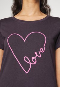 Mörkgrå T-shirt med en stor, ljusrosa hjärtoutline där ordet "kärlek" är integrerat i designen. Kort ärm, rund halsringning.
