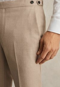 Bežiniai tekstūruoti džinsai su dviem juodais mygtukais juosmens srityje, turintys šoninį kišenę ir pritaikytą siluetą.