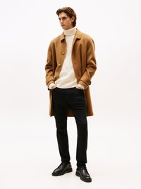Abrigo de lana color camello con grandes botones sobre un cuello de tortuga de punto blanco, combinado con pantalones negros y zapatos negros de cordones. Diseño simple y moderno.