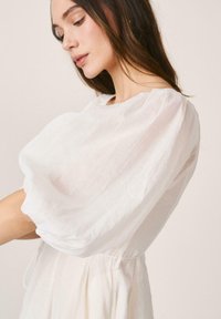 Mujer con cabello largo y oscuro, vestida con un vestido blanco de mangas abullonadas y voluminosas, mirando hacia abajo contra un fondo liso.