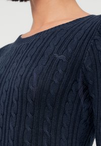 Πλεκτό πουλόβερ σε ναυτικό μπλε, με σχέδιο καλώδιο, στρογγυλή λαιμόκοψη και μακριά μανίκια, που αναδεικνύει υφή ύφανσης.