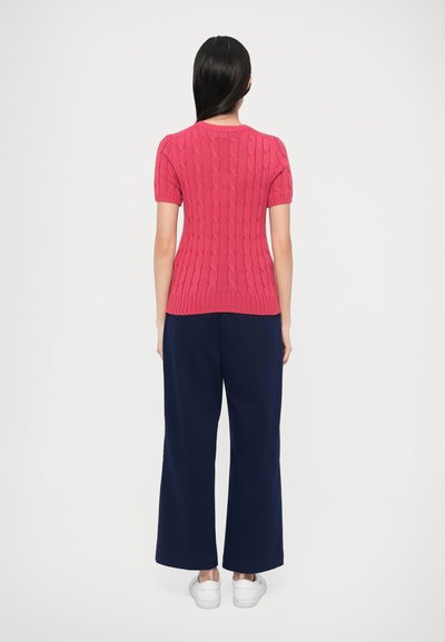 Φόρεμα με κοντά μανίκια, εφαρμοστό, πλεκτό με σχέδιο κοτσίδες σε ζωηρό ροζ χρώμα, σε συνδυασμό με φαρδιά, χαλαρά παντελόνια σε ναυτικό μπλε και λευκά αθλητικά παπούτσια.