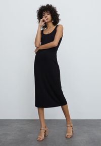 Zwarte ribbel midi-jurk met een aansluitende silhouet en zonder mouwen. Gecombineerd met tan blokhak sandalen, tegen een neutrale achtergrond.