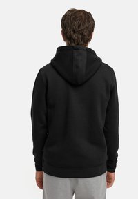 Svart hettegenser med en jevnt tekstur, ribbede mansjetter og kengurulomme, iført med grå joggebukser sett fra bak. 