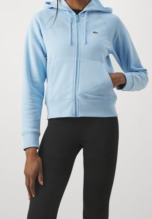 Persoon die een lichtblauwe hoodie met rits en zakken draagt en zwarte aansluitende legging, hand in de zak, tegen een effen lichte achtergrond.