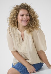 Beige, short-sleeve blouse with a v-neck, featuring vertical pleats and lace detail at the neckline, paired with blue striped shorts.