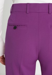 Lila skräddarsydda byxor i ett strukturerat tyg, med två bakfickor med svarta knappar och en platt midja.