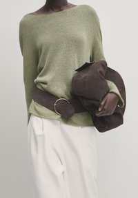 Maglione verde lavorato a maglia con design off-shoulder, abbinato a una cintura in suede marrone scuro con fibbia circolare; pantaloni bianchi su misura.