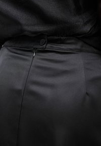 Κοντινή λήψη μαύρου ραμμένου παντελονιού με πλευρικό φερμουάρ και κλείσιμο με κουμπί, φορεμένο με μαύρο τοπ που έχει μπει μέσα.