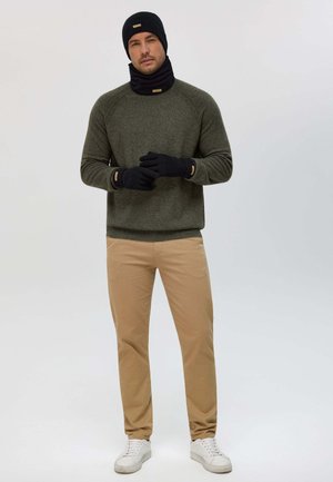 Grüner Pullover, schwarze Strickmütze und Schal, schwarze Handschuhe, beige Hose und weiße Sneakers. Der Pullover hat gerippte Bündchen und Struktur.