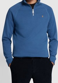 Pull-over bleu à demi-fermeture éclair en tissu doux avec poignets côtelés, présentant un petit logo brodé sur la poitrine.