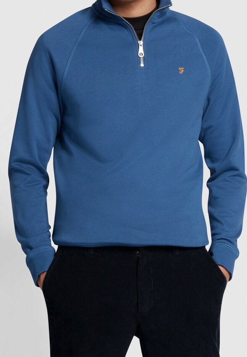 Pull-over bleu à demi-fermeture éclair en tissu doux avec poignets côtelés, présentant un petit logo brodé sur la poitrine.