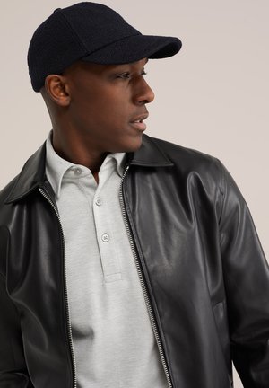 Hombre con chaqueta de cuero negra, camisa gris claro con cuello y gorra negra texturizada, mirando hacia un lado sobre un fondo liso.